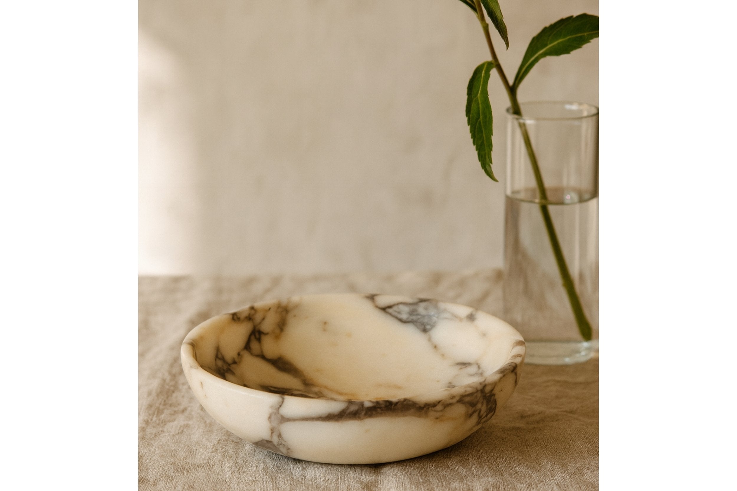 Calacatta Viola marble bowl styled on linen table with glass vase and soft natural light, minimal home decor scene.