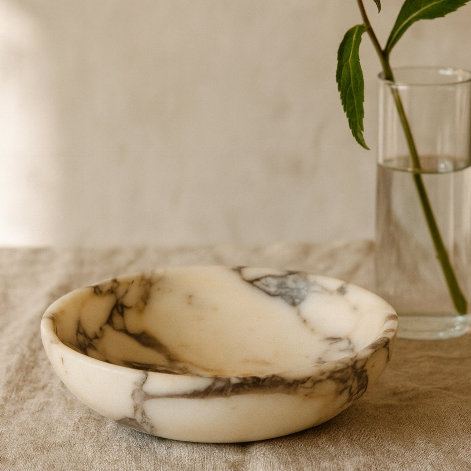 Calacatta Viola marble bowl styled on linen table with glass vase and soft natural light, minimal home decor scene.