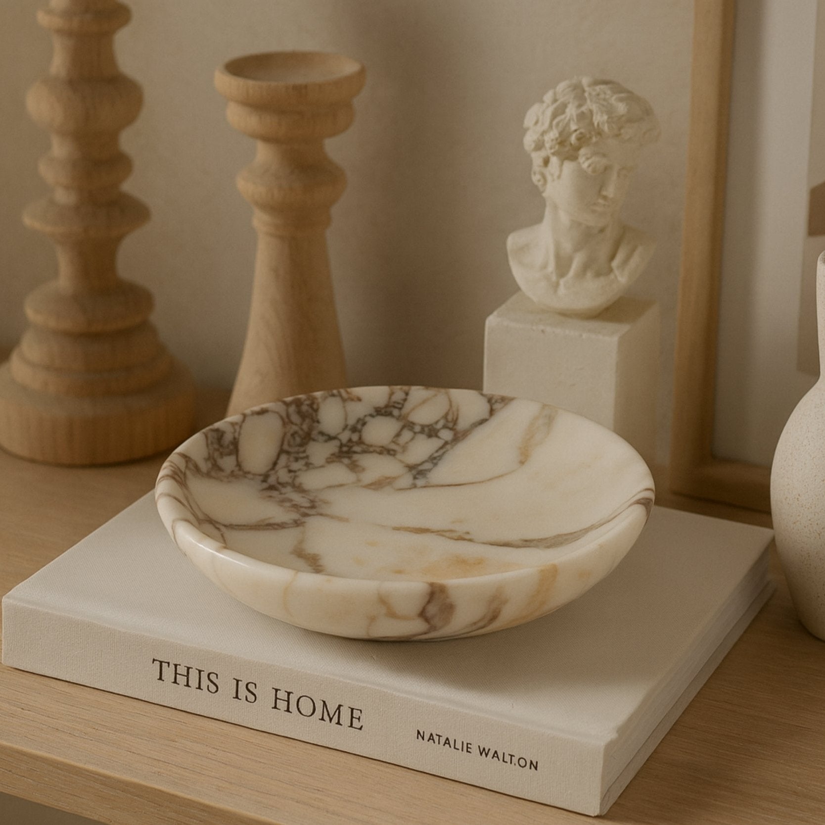 Calacatta Viola marble plate on “This Is Home” book surrounded by wooden candlesticks and art sculpture, reflecting timeless elegance.