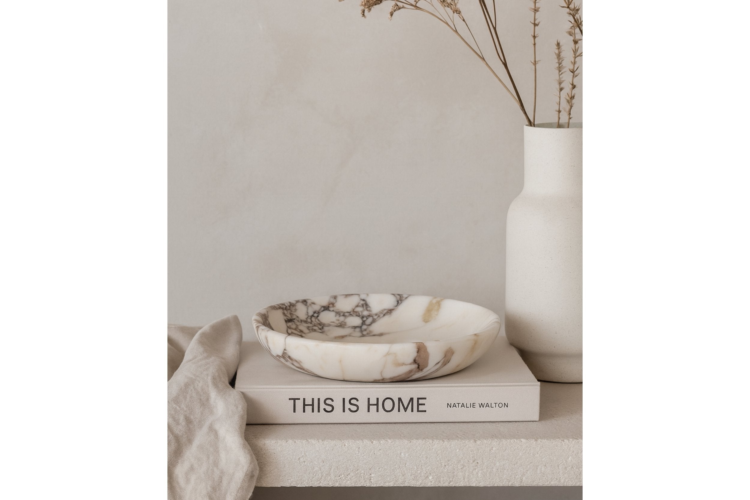 Minimalist scene featuring Calacatta Viola marble plate on neutral books with soft shadows and natural dried florals.