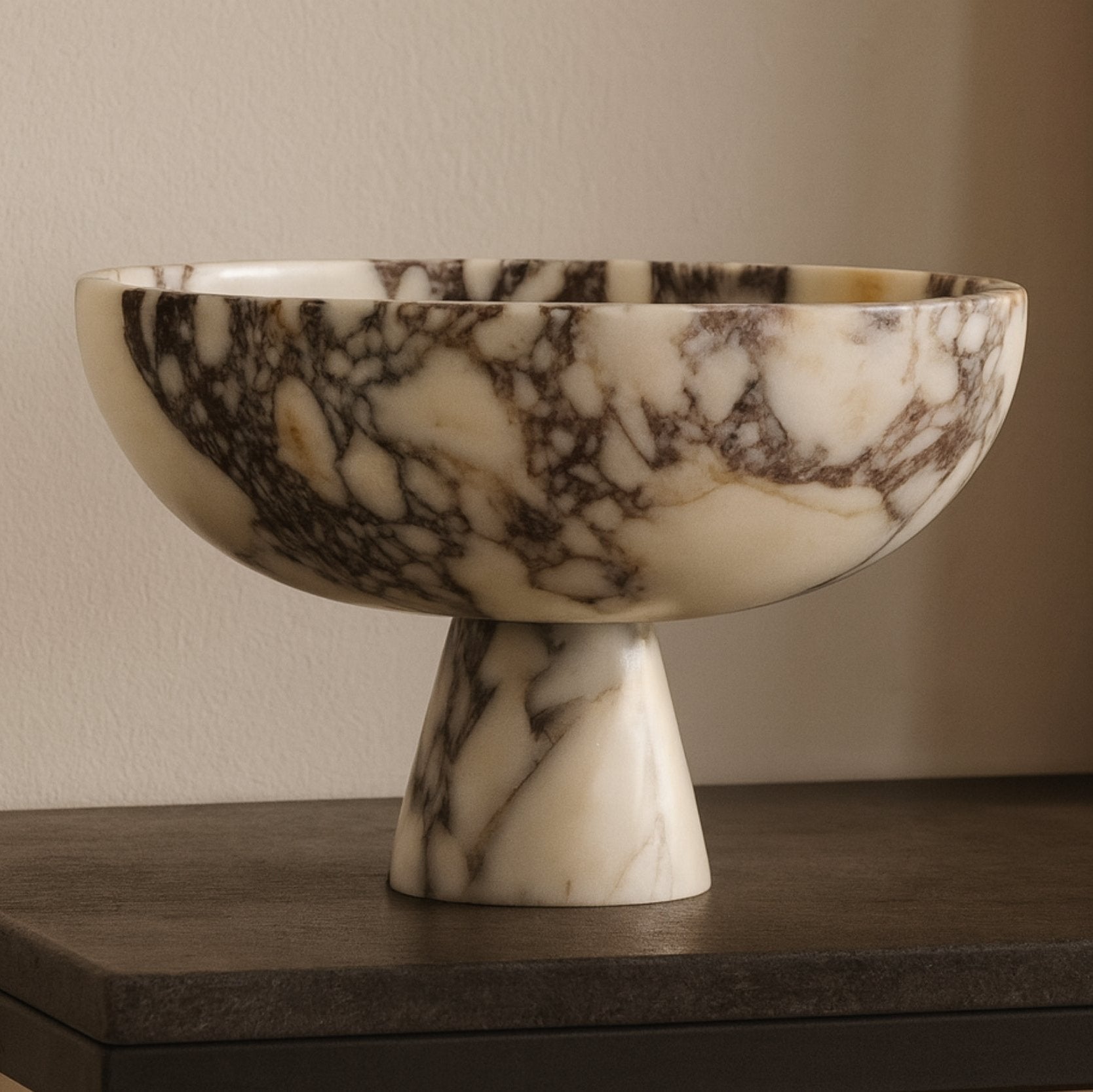 calacatta viola marble pedestal bowl styled on neutral shelf with books and sculptures, modern interior soft daylight
