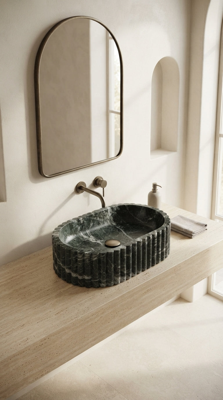 Oval fluted marble sink on a pale stone vanity in a bright bathroom with an arched mirror and brass wall-mounted faucet.