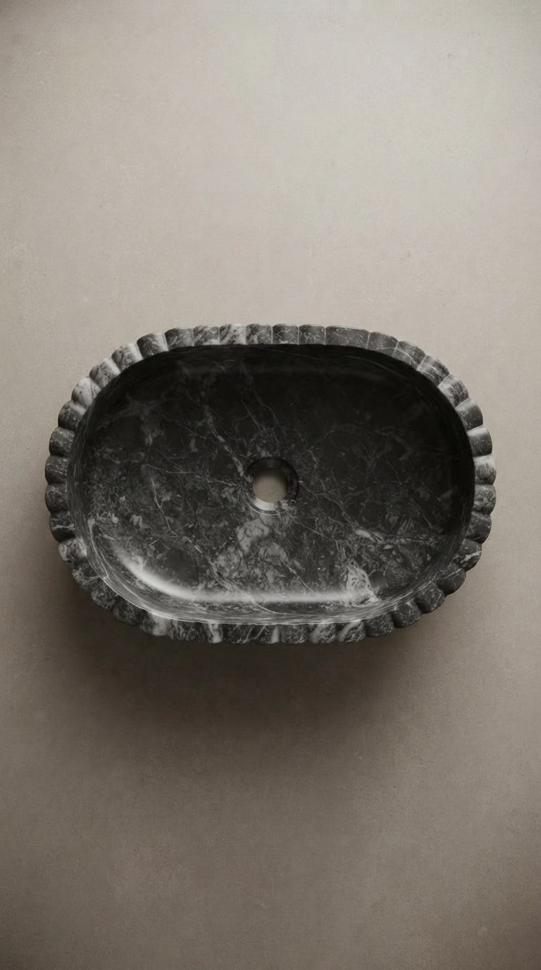 Overhead view of an oval marble vessel sink showing its natural veining, curved basin, scalloped outer edge and drain opening.